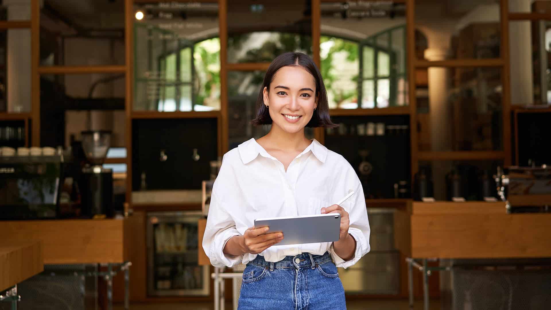 A small cafe owner smiling and holding an iPad in her cafe, showcasing entrepreneurship and business growth.