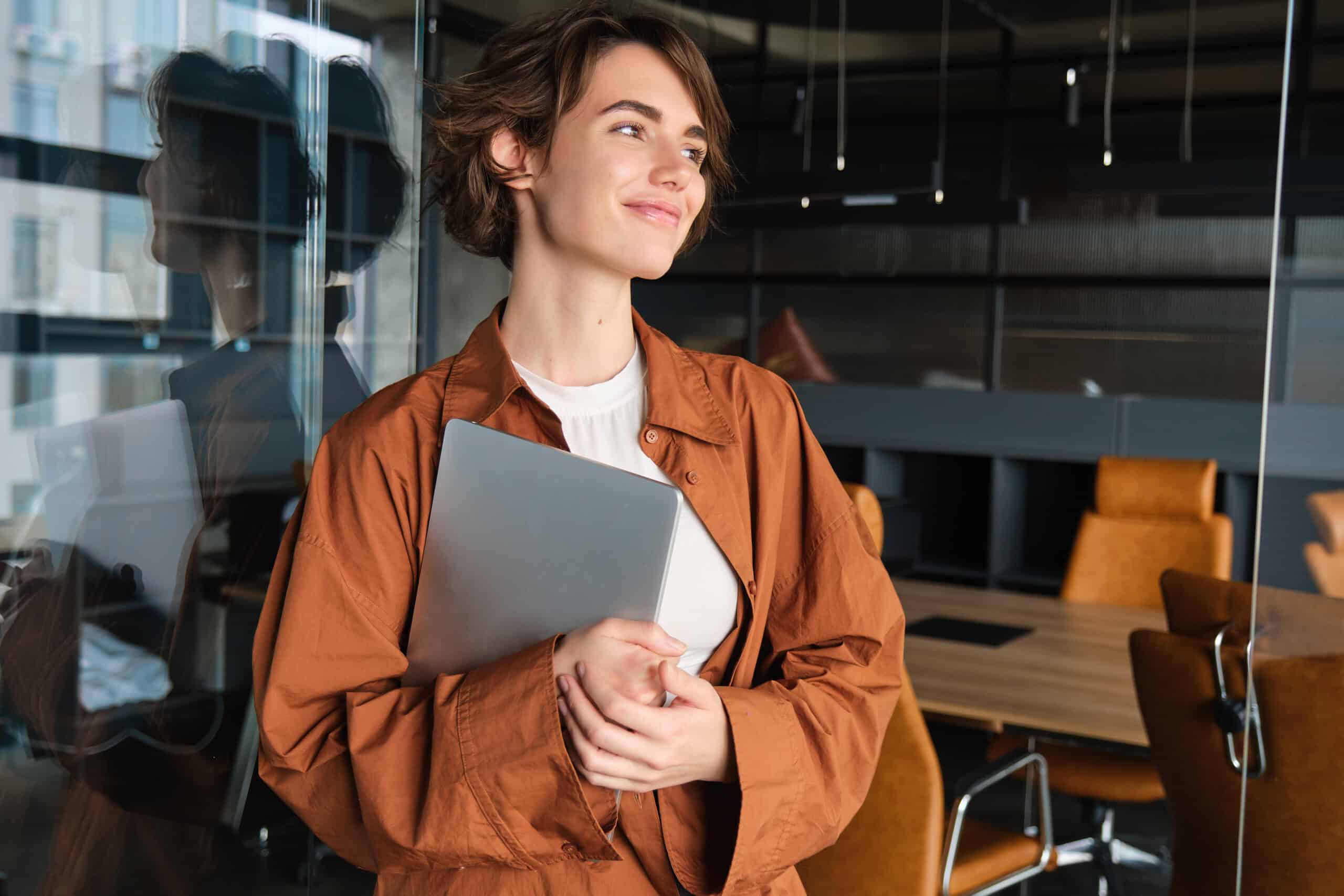 Professional woman holding a laptop and smiling in a modern office environment.